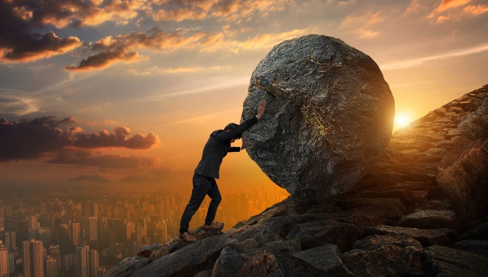 A businessman laboriously pushing a rock uphill.