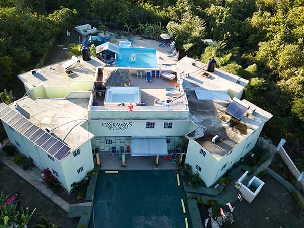 An aerial view of Castaway's Villa, a rectangular resort with a rooftop pool and lounging area. The villa is surrounded by lush greenery and features multiple solar panels on the roof. Flags are displayed near the entrance, making it an ideal venue for small business conferences amidst nature.