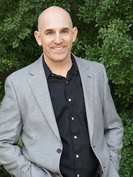 Brian Gibbs is smiling while posing with his hands in his pockets. He is wearing a black shirt and a gray plaid blazer, embodying the essence of smart business style. Behind him, the dense green foliage adds a refreshing touch to this Smart Business Accelerator promotional shoot.