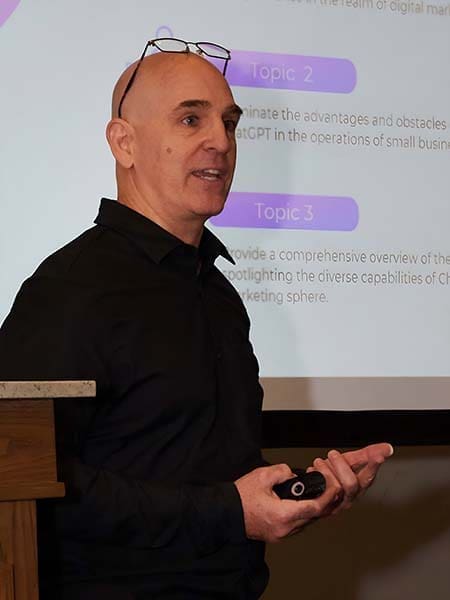 Brian Gibbs, wearing a black shirt and glasses on his head, is giving a presentation on small business digital marketing. Holding a small device in his hand, he stands in front of a projected screen showing text with headings "Topic 2" and "Topic 3," beside a wooden podium. Brian Gibbs, wearing a black shirt and glasses on his head, is giving a presentation on small business digital marketing. Holding a small device in his hand, he stands in front of a projected screen showing text with headings "Topic 2" and "Topic 3," beside a wooden podium.