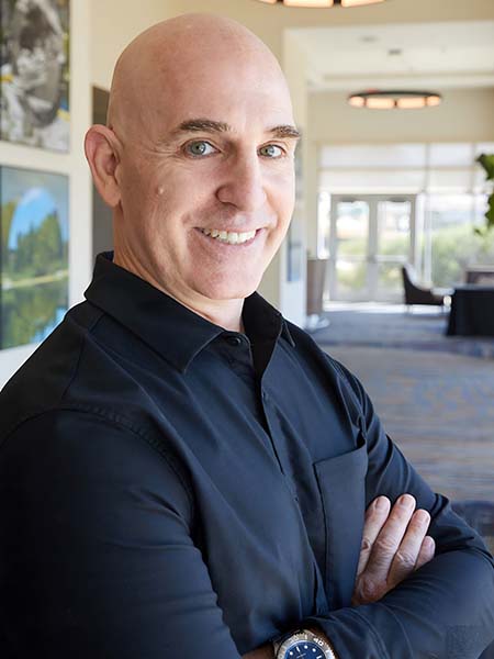 Brian Gibbs, a smiling bald man in a dark blue shirt, stands with his arms crossed in a well-lit, modern office hallway. Framed pictures adorn the walls, and large windows flood the space with natural light. Brian specializes in small business digital marketing and is a key player in digital marketing San Antonio.