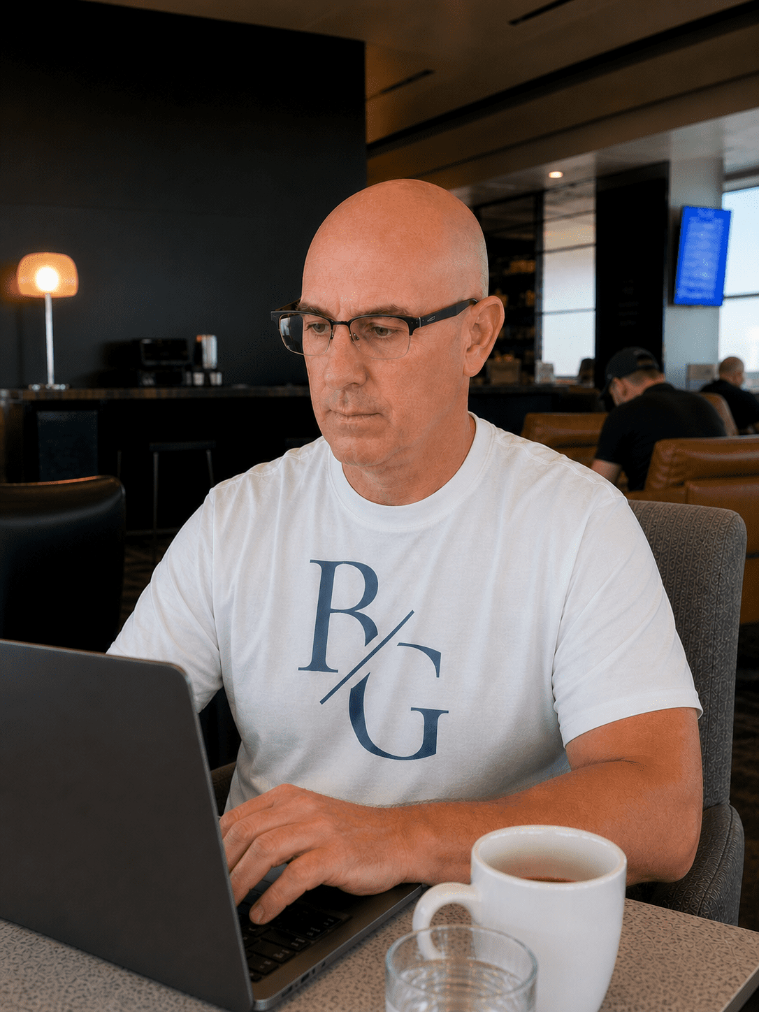Brian Gibbs working on a laptop in an airport lounge, wearing a white BG monogram t-shirt with a coffee cup on the table