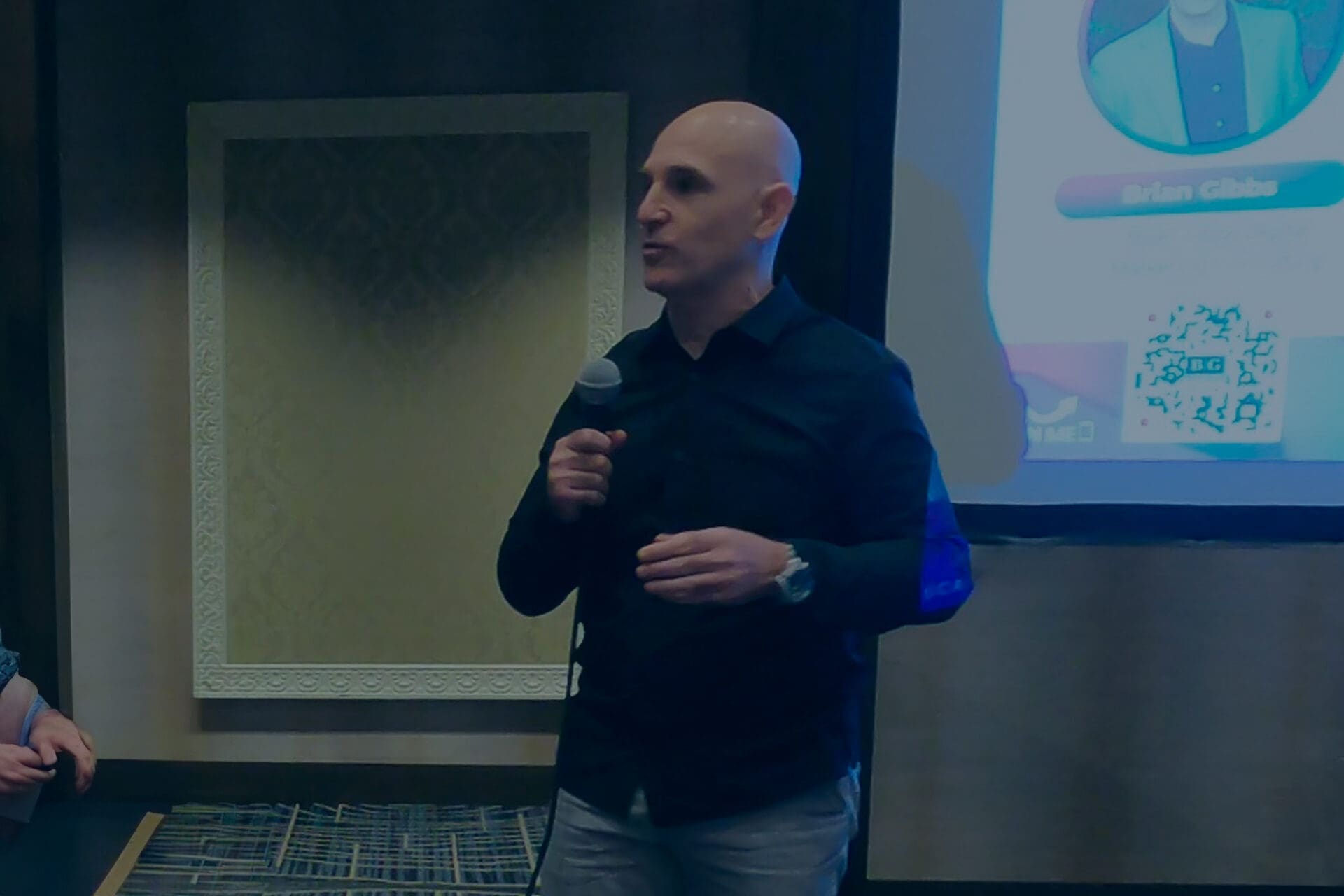 Brian Gibbs wearing a dark shirt discusses AI strategies for SMBs into a microphone while standing in front of a projected presentation screen and a decorative frame on the wall.