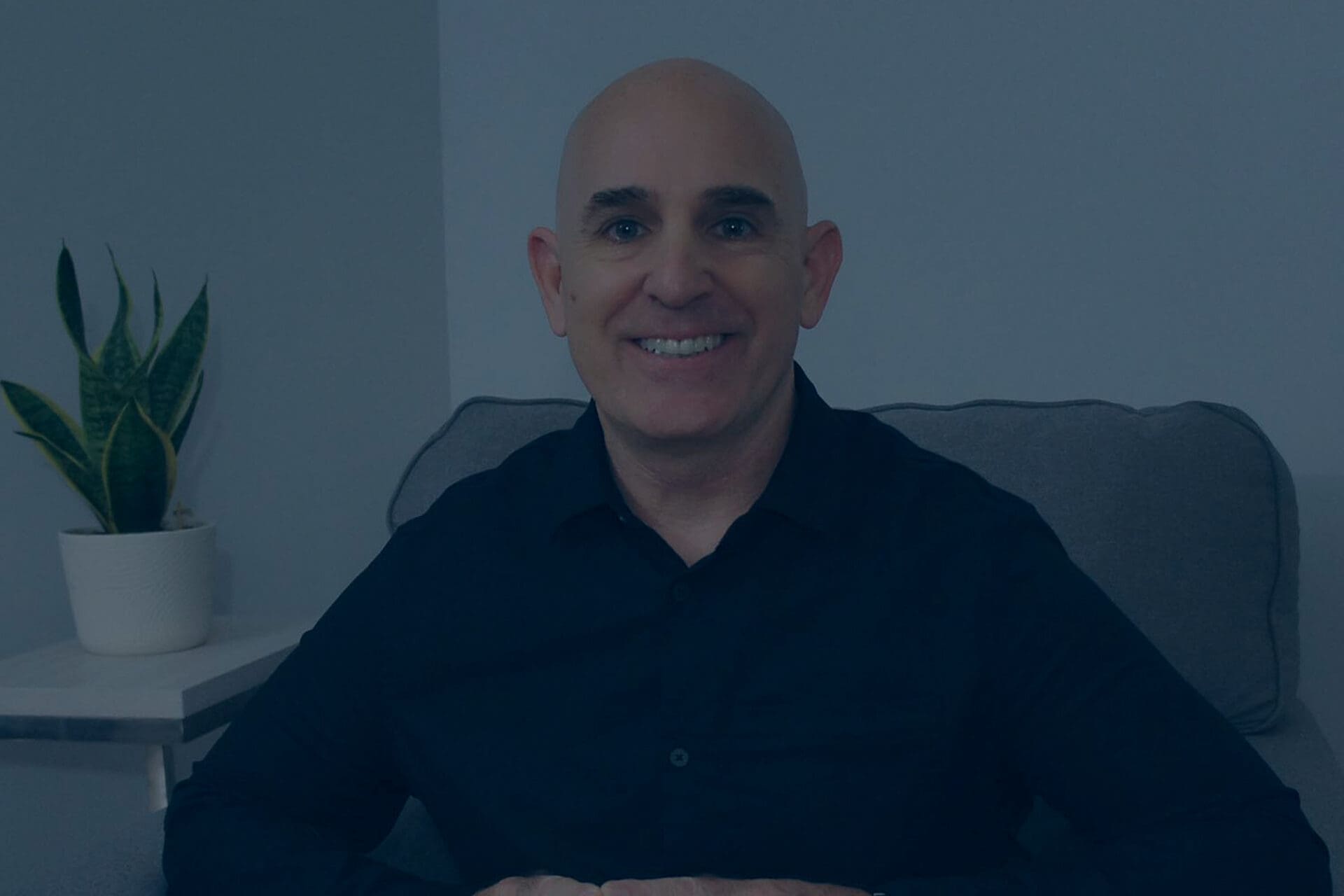 Brian in a dark shirt sits on a gray chair indoors, discussing AI strategies for SMBs, with a green potted plant on a small table next to him.