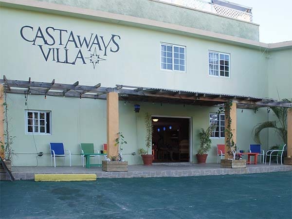 Front view of Castaways Villa, a pale green building with a wooden pergola. The entrance is adorned with potted plants and colorful chairs under a shaded patio. The name is prominently displayed on the upper left side of the building.