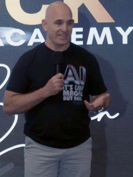 Brian Gibbs in a black T-shirt with colorful text holds a microphone while speaking. He stands in front of a backdrop with partially visible white lettering. His expression is friendly and engaged.