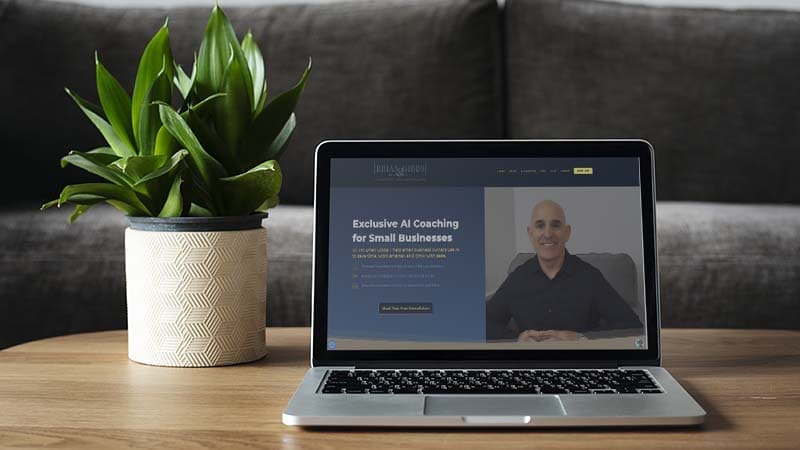 A laptop on a wooden table shows a website with a man smiling and text about exclusive AI coaching for small businesses. A green plant in a patterned pot is placed next to the laptop.