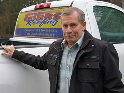 My dad, Doug Gibbs, wearing a plaid shirt and dark jacket stands beside a white truck with "Gibbs Roofing" on the rear window. The background shows trees and an overcast sky.