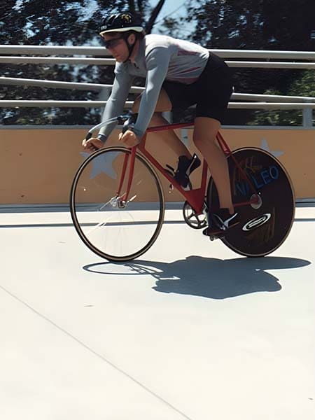 A cyclist wearing a helmet rides a red track bike on an outdoor velodrome. The blurred background indicates speed, with sunlight casting shadows on the ground.