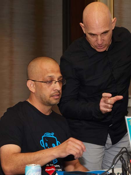 Brian interacting at a table with a laptop. One man is seated, wearing glasses and a black t-shirt, while the other stands beside him, wearing a black long-sleeve shirt, gesturing as he talks.