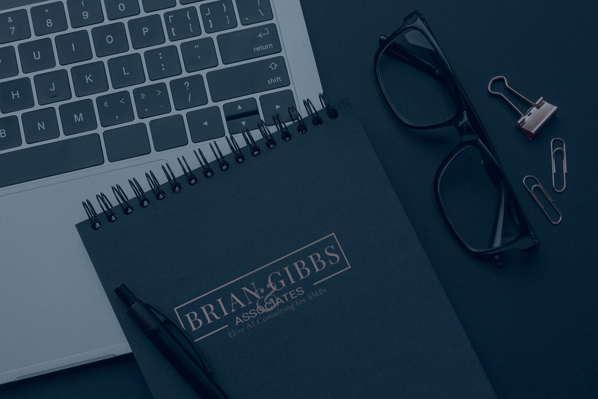 A close-up of a laptop keyboard, black-rimmed eyeglasses, a spiral notebook labeled "Brian Gibbs & Associates," a pen, paper clips, and a binder clip arranged on a dark surface—ideal tools for SMBs developing effective AI strategies.