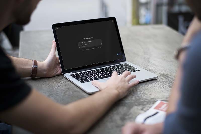 Two people are sitting at a table, looking at a MacBook laptop. One person is using the trackpad. The screen displays a dark interface with a form asking "What’s on your mind?" in white text. A book is on the table beside them.