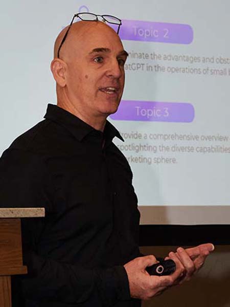 Brian Gibbs is standing in front of a presentation screen, speaking and holding a remote control. They are wearing glasses and a dark shirt. The slide behind highlights topic points related to business applications of technology.