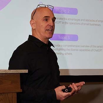 Brian Gibbs is standing in front of a presentation screen Brian Gibbs is standing in front of a presentation screen, speaking and holding a remote control. They are wearing glasses and a dark shirt. The slide behind highlights topic points related to business applications of technology.