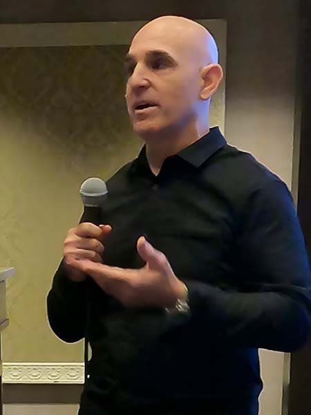 Brian Gibbs speaks into a microphone, gesturing with his other hand. He stands indoors against a blurred decorative background.