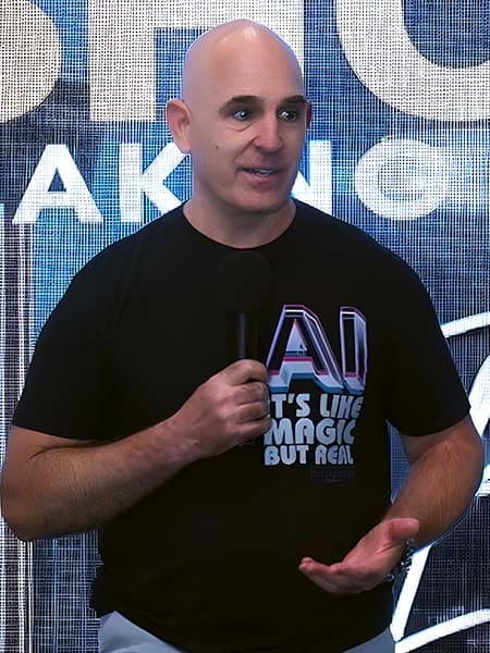 A person with a shaved head is speaking into a microphone while wearing a black T-shirt with "AI, it's like magic but real" printed on it. Behind them is a large illuminated sign.