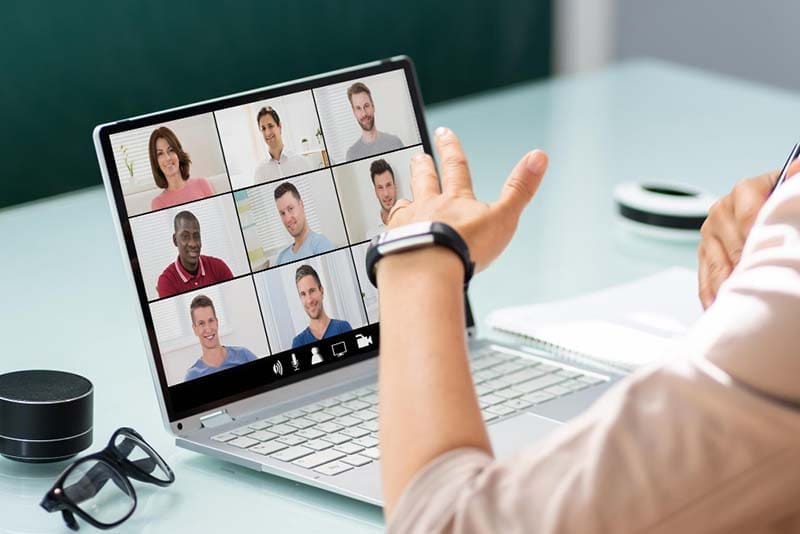 A person sitting at a desk participates in a video conference call on a laptop. The screen displays eight diverse individuals in separate video boxes. Nearby are a notebook, pen, smartwatch, glasses, and a speaker.